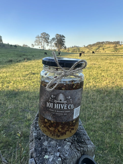 101 Hive Co. Hot Honey 500g jar with chilli pieces visible, sitting on a rustic fence post with cattle grazing in the background
